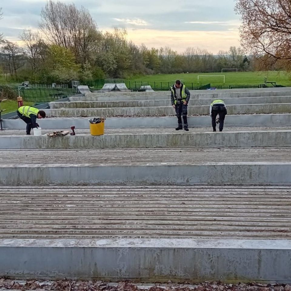 Three workers in high-visibility vests are cleaning or inspecting concrete steps in an outdoor setting with trees and a sports field in the background. Three workers in high-visibility vests are cleaning or inspecting concrete steps in an outdoor setting with trees and a sports field in the background.